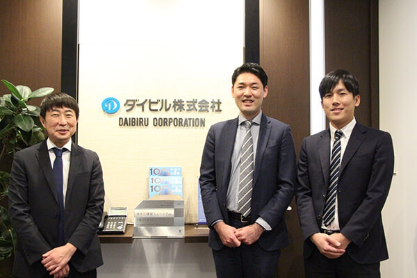 ダイビル株式会社のロゴ前で撮影された担当者3名の写真
